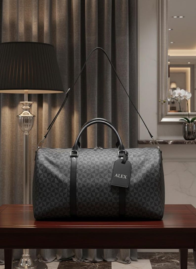 A gray patterned duffel bag with a black handle and tag is placed on a wooden table in a room with a lamp and a mirror.