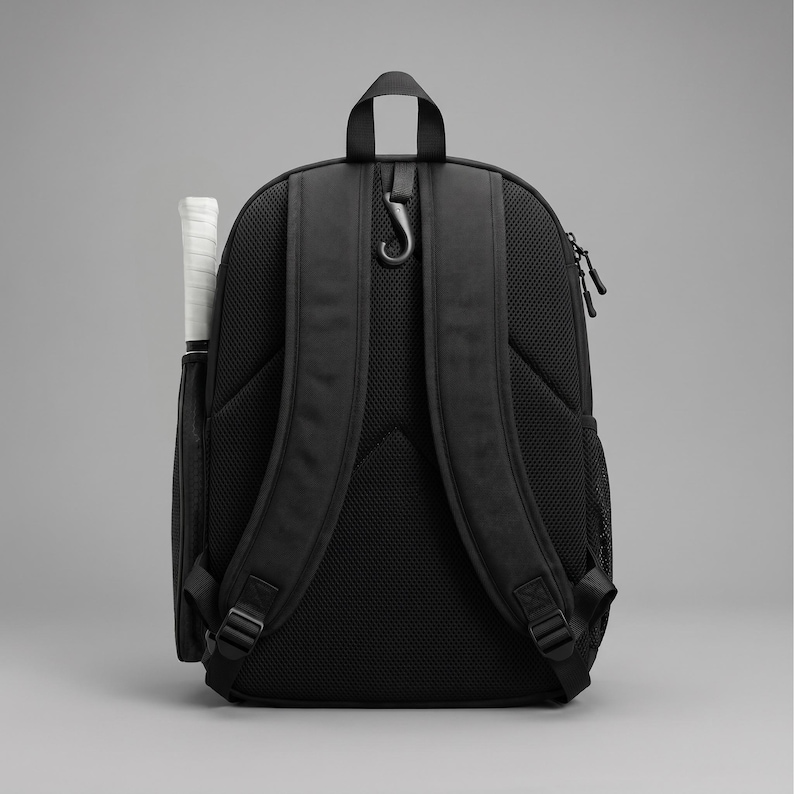 May include: A black backpack featuring padded shoulder straps with a mesh back panel, a top carry handle, and a black hook attachment. A white-handled racket is stored in a side pocket, and a black mesh pocket is visible on the opposite side.