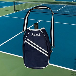 May include: A navy blue tennis racket bag with a white strap and white accents. The bag has a white embroidered name "Sarah" on the front.