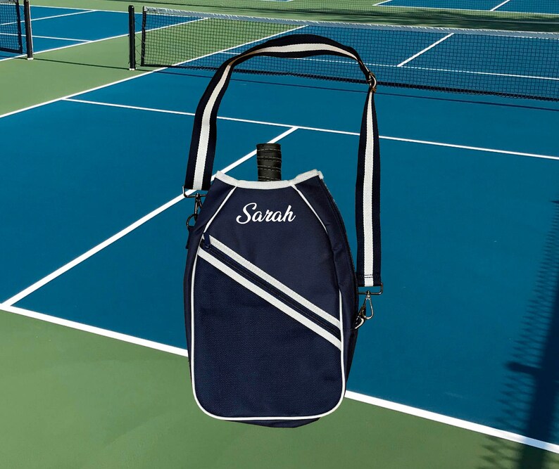 May include: A navy blue tennis racket bag with a white strap and white accents. The bag has a white embroidered name "Sarah" on the front.