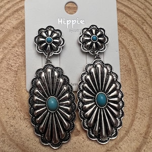 May include: A pair of silver-toned metal drop earrings featuring a flower-shaped stud and a large oval pendant. Both components are decorated with a central turquoise-colored cabochon and radial grooved detailing. The earrings are attached to a white display card with the text Hippie printed at the top.