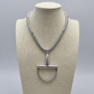 May include: A silver-toned necklace featuring a flexible, coiled metal chain that connects to a large, polished metal bit pendant. The pendant is shaped like a horse bit with a central vertical bar and a rounded, semi-circular lower loop.