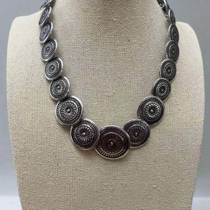 May include: A silver-toned necklace displayed on a mannequin. The necklace features a series of circular, embossed pendants with a floral design. The pendants are linked together to form a statement piece of jewelry.