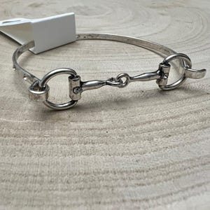 May include: A silver-toned metal bangle bracelet featuring a horse bit design. The bracelet has a smooth, polished finish and includes small, clear embedded crystals along the band and the central bit component, with a white tag attached.