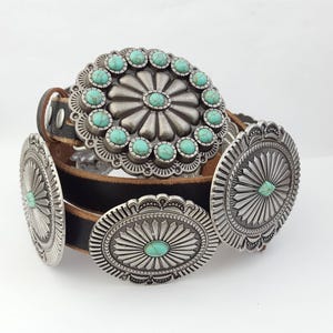 May include: A black leather belt featuring large, circular silver-toned metal conchos. The central concho is adorned with a ring of small, round turquoise-colored stones, while the other conchos feature a single central turquoise-colored stone.