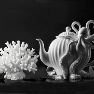 May include: A white ceramic teapot with an octopus wrapped around it. The teapot has a round body and a spout. The octopus has eight tentacles and is curled around the teapot. There is a white coral reef to the left of the teapot.