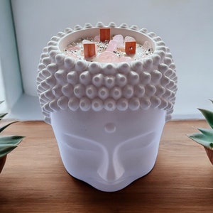 Puede incluir: Portavelas de cerámica blanca con forma de cabeza de Buda con una vela rosa y blanca en su interior. La vela está decorada con pequeños cristales rosas y blancos y palitos de madera.
