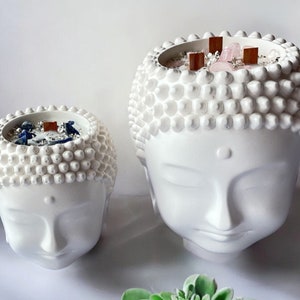 Puede incluir: Dos portavelas de cerámica blanca en forma de cabeza de Buda con una superficie texturizada. El portavelas más grande tiene una vela rosa con cristales y palitos de madera en su interior. El portavelas más pequeño tiene una vela azul con cristales y palitos de madera en su interior.