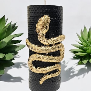 May include: A black pillar candle with a textured surface, adorned with a gold-colored snake wrapped around it. The snake has a textured, metallic appearance. A white wick sits atop the candle. The candle is a decorative item.