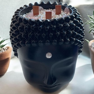May include: Black Buddha head candle holder with a white dot on the forehead. The holder is filled with pink and clear crystals and wooden wicks.