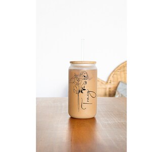 May include: A clear glass jar with a bamboo lid and a straw. The jar is filled with a brown liquid and has a black and white floral design with the word "Tracy" written on it.