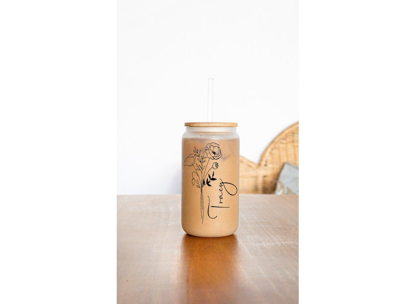 May include: A clear glass jar with a bamboo lid and a straw. The jar is filled with a brown liquid and has a black and white floral design with the word "Tracy" written on it.