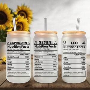 Op de afbeelding: Drie frosted glazen bekers met bamboe deksels en rietjes. Elke beker heeft een ander sterrenbeeld: Steenbok, Tweelingen en Leeuw. De bekers hebben een witte achtergrond met zwarte tekst die "Nutrition Facts" luidt en verschillende persoonlijkheidstrekken met een percentagewaarde vermeldt.