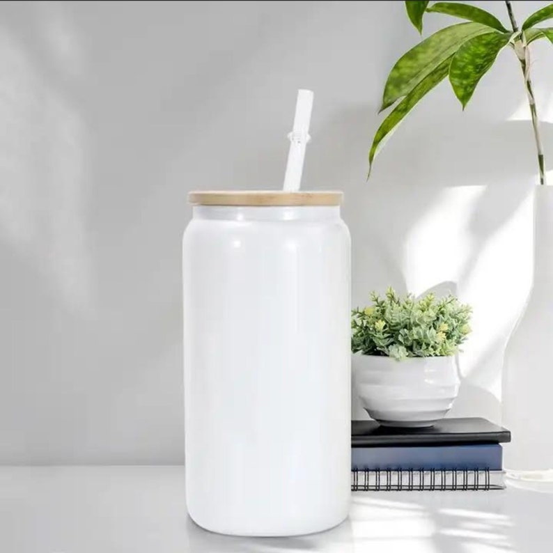 Op de afbeelding: Witte keramische blik met een houten deksel en een witte rietje die uit het deksel steekt. De blik staat op een wit oppervlak.