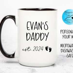 May include: White ceramic mug with a black handle and rim. The mug has black text that reads "Evan's Daddy est. 2024" with two small black footprints.