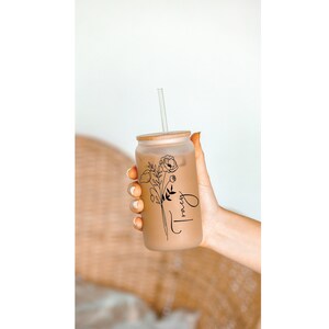 May include: A clear glass tumbler with a brown lid and a black floral design. The tumbler has a straw and says "Tracy" on the side.