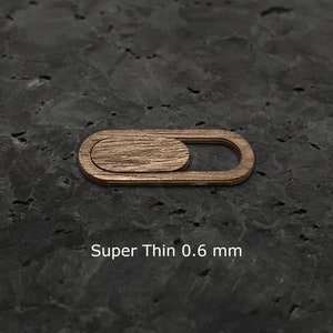 May include: A thin, wooden webcam cover in a light brown color. The cover is designed to slide over the webcam lens to protect privacy. The text "Super Thin 0.6 mm" is printed below the cover.