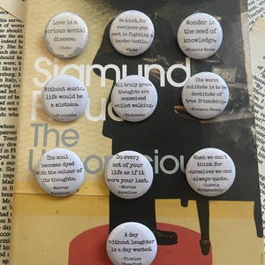 Könnte beinhalten: Zehn runde, weiße Ansteckbuttons mit schwarz gedruckten philosophischen Zitaten von Autoren wie Platon, Nietzsche, Francis Bacon, Marc Aurel, Ludwig Wittgenstein und Nicolas Chamfort, die in einem Raster auf einem Buchumschlag angeordnet sind.