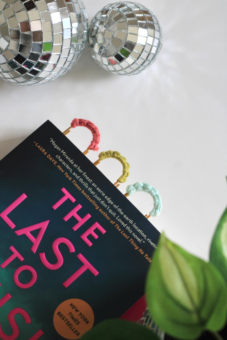 以下が含まれることがあります： ピンク色の文字で「The Last to Us」と書かれた本と、3つのカラフルな手作りのブックマーク。ブックマークはピンク、黄緑、水色です。背景には2つのディスコボールがあります。