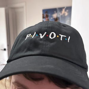Puede incluir: Gorra de béisbol negra con la palabra "PIVOT!" bordada en blanco, con puntos de colores sobre las letras. La gorra tiene una visera curva y está hecha de una tela suave.