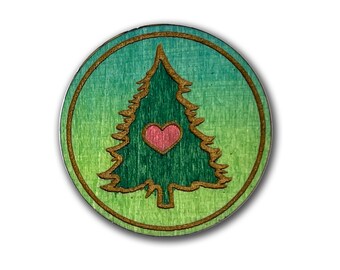 May include: A round wooden ornament with a green and blue gradient background. A brown outline of a pine tree with a pink heart in the center is in the middle of the ornament.