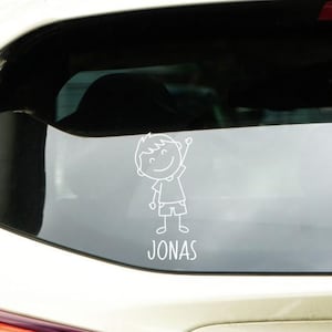 Op de afbeelding: Witte autosticker met een eenvoudige, lachende cartoonjongen die zwaait. De naam "JONAS" staat eronder. De sticker is op de achterruit van een witte auto geplakt.