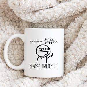 May include: White ceramic coffee mug with a black graphic of a stick figure giving the middle finger. The mug has the German text "VOR DEM ERSTEN Kaffee KLAPPE HALTEN !!!" printed on it.