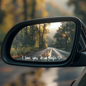 Può includere: Specchietto retrovisore di un'auto che riflette una strada attraverso una foresta. Il testo "I love you, drive safe" è scritto in bianco nella parte inferiore dello specchietto.