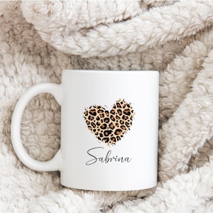 Könnte beinhalten: Weiße Keramik-Tasse mit einem Leopardenmuster-Herz-Design und dem Namen "Sabrina" in Schreibschrift. Die Tasse hat einen gebogenen Henkel. Der Hintergrund ist ein weiches, strukturiertes Gewebe.