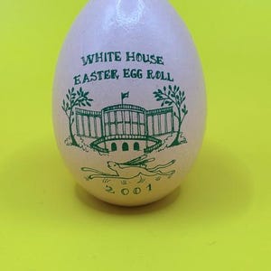 Puede incluir: Un huevo coleccionable de color rosa claro con texto y gráficos verdes. El texto dice "WHITE HOUSE EASTER EGG ROLL" encima de una representación de la Casa Blanca y un conejo corriendo. El año "2001" está impreso debajo.