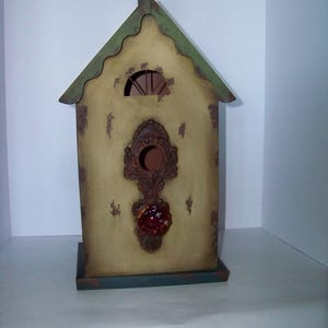 Könnte beinhalten: Ein dekoratives Vogelhaus mit einem Used-Look-Finish. Das Vogelhaus hat einen hellgelben Körper, ein grünes Dach und verzierte, dunkelbraune Verzierungen. Ein roter, juwelenartiger Akzent befindet sich mittig auf der Vorderseite. Das Vogelhaus hat ein kleines, gewölbtes Fenster und eine dekorative Spitze.