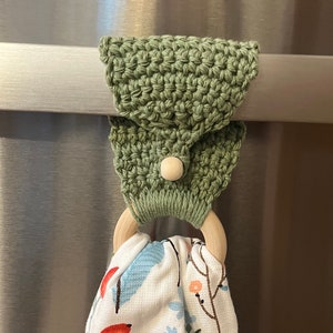 以下が含まれることがあります： 白いボタンと木製リングが付いた緑色のクロシェタオルリング。リングはステンレス製の表面に掛けられており、白と青の花柄のタオルがリングに掛けられています。