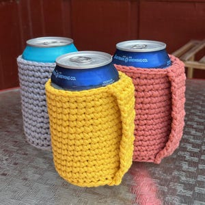 May include: Three crocheted can coolers in gray, yellow, and coral. The coolers are made with a chunky yarn and have a simple, modern design.