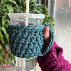 May include: A teal crocheted cup cosy with a handle, holding a clear plastic cup with a straw and water.