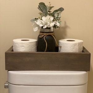 May include: A wooden tray with two rolls of toilet paper and a black mason jar with a floral arrangement. The tray is sitting on top of a white toilet tank.