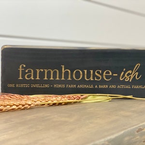 Puede incluir: Un letrero de madera negro con letras doradas que dice "farmhouse-ish" y "ONE RUSTIC DWELLING - MINUS FARM ANIMALS, A BARN AND ACTUAL FARMLAND".