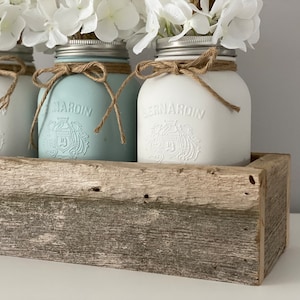 Op de afbeelding: Vier witte geverfde glazen potten met een blauw-wit ontwerp, vastgebonden met touw en gevuld met witte bloemen, die in een rustieke houten plantenbak staan.