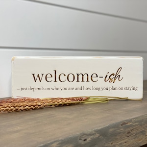 May include: White wooden sign with the text "welcome-ish... just depends on who you are and how long you plan on staying" in gold lettering.