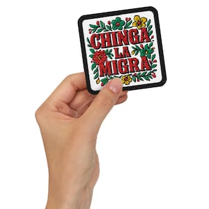 Peut inclure: Écusson carré brodé avec le texte "CHINGA LA MIGRA" en rouge, entouré de motifs floraux verts, rouges et jaunes. L'écusson a une bordure noire et est tenu dans une main.