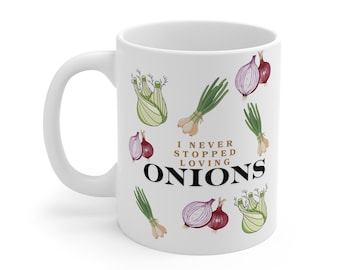 May include: A white ceramic mug featuring a repeating pattern of various onions and fennel bulbs in shades of purple, green, and white. The text "I NEVER STOPPED LOVING ONIONS" is printed in the center in a combination of brown and black fonts.