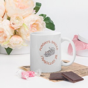 Peut inclure: Mug en céramique blanche avec une anse, orné d'une illustration de chat gris et du texte "MONDAYS SHOULD BE OPTIONAL!". La tasse est entourée de fleurs roses, de bonbons emballés et de chocolat.