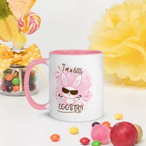 Könnte beinhalten: Weißer Keramikbecher mit rosa Innenraum und Griff. Der Becher zeigt einen Cartoon-Hasen mit Sonnenbrille und den Text "I'm a little EGGSTRA" auf einem rosa Aquarellhintergrund. Bunte Süßigkeiten und Dekorationen umgeben den Becher.
