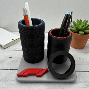 May include: A desk tidy with pen holders designed as stacked black tyres with blue and red accents. The organiser includes a grey base with a red F1 logo and holds pens and a small succulent in a terracotta pot.