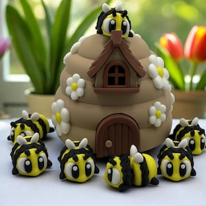 May include: A charming, handmade bee-themed decorative set. The focal point is a tan beehive with a brown door and window, decorated with white daisy accents. Several yellow and black bee figurines surround the hive, creating a delightful, playful scene.
