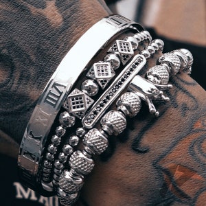 May include: A set of silver bracelets with various designs, including a beaded bracelet, a bracelet with black stones, and a bracelet with Roman numeral markings.