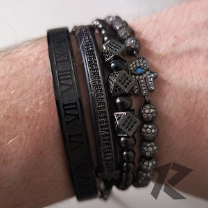 Puede incluir: Un conjunto de pulseras negras con números romanos, cuentas y dijes de mano de hamsa. Las pulseras están apiladas en la muñeca de una persona.