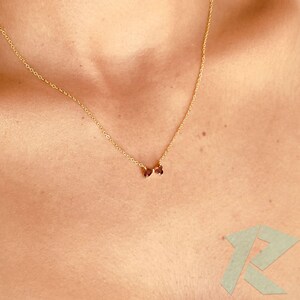 May include: A fine gold necklace with two small, oval, dark red gemstones. The necklace is worn on a person's chest, highlighting the delicate chain and the subtle sparkle of the gemstones.