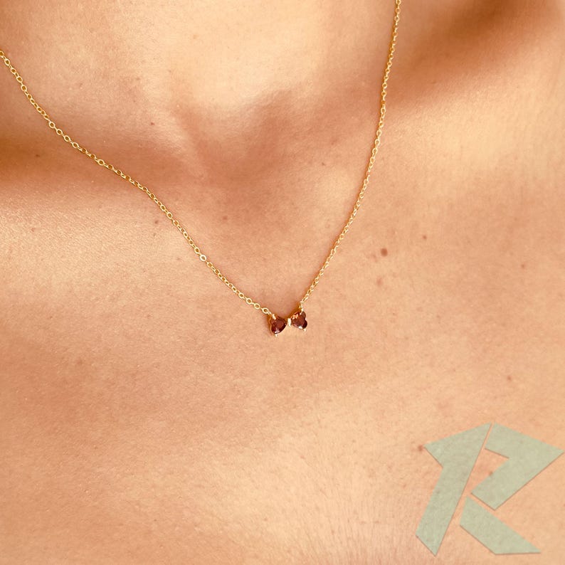 May include: A fine gold necklace with two small, oval, dark red gemstones. The necklace is worn on a person's chest, highlighting the delicate chain and the subtle sparkle of the gemstones.
