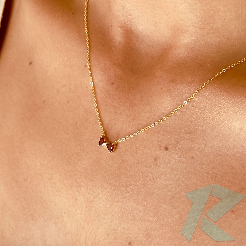 May include: A delicate gold necklace with a small pendant featuring two heart-shaped, reddish-brown gemstones. The necklace has a fine chain and is displayed against a skin-toned background. The pendant is centered, creating a minimalist and elegant design.