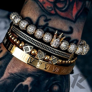 May include: A selection of gold and beaded bracelets, some with sparkling cubic zirconia stones. One bracelet displays Roman numerals. A gold crown charm is also visible. The bracelets are stacked together.